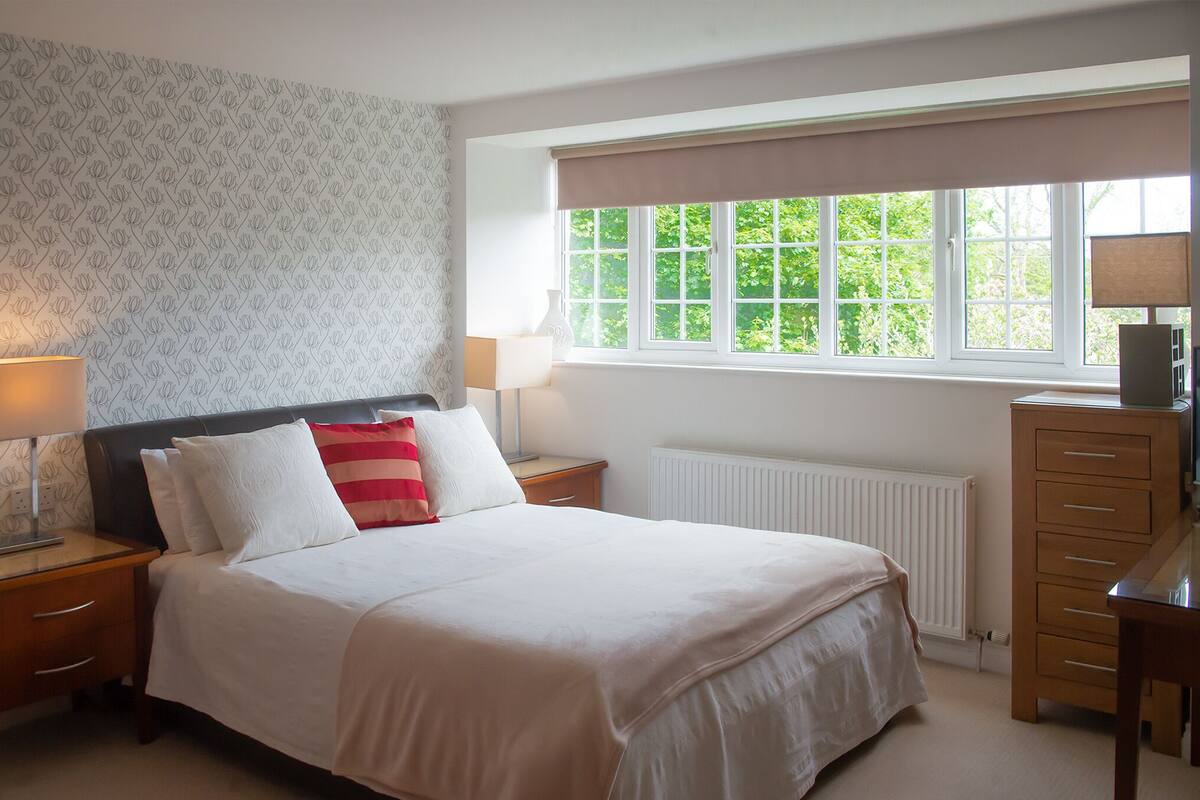 A bedroom with a bed, two bedside tables, a lamp, a dresser, and a window with a view of greenery.