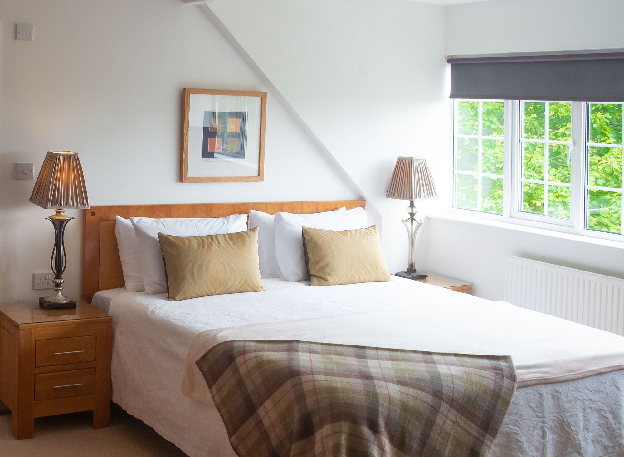 A bedroom with a bed, two bedside tables, lamps, a framed picture, and a window with a view of greenery.