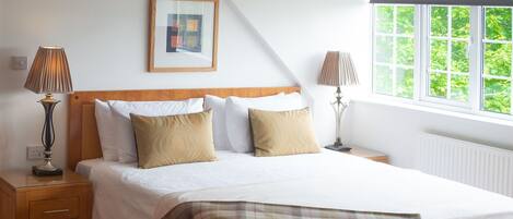 A bedroom with a bed, two bedside tables, lamps, a framed picture, and a window with a view of greenery.