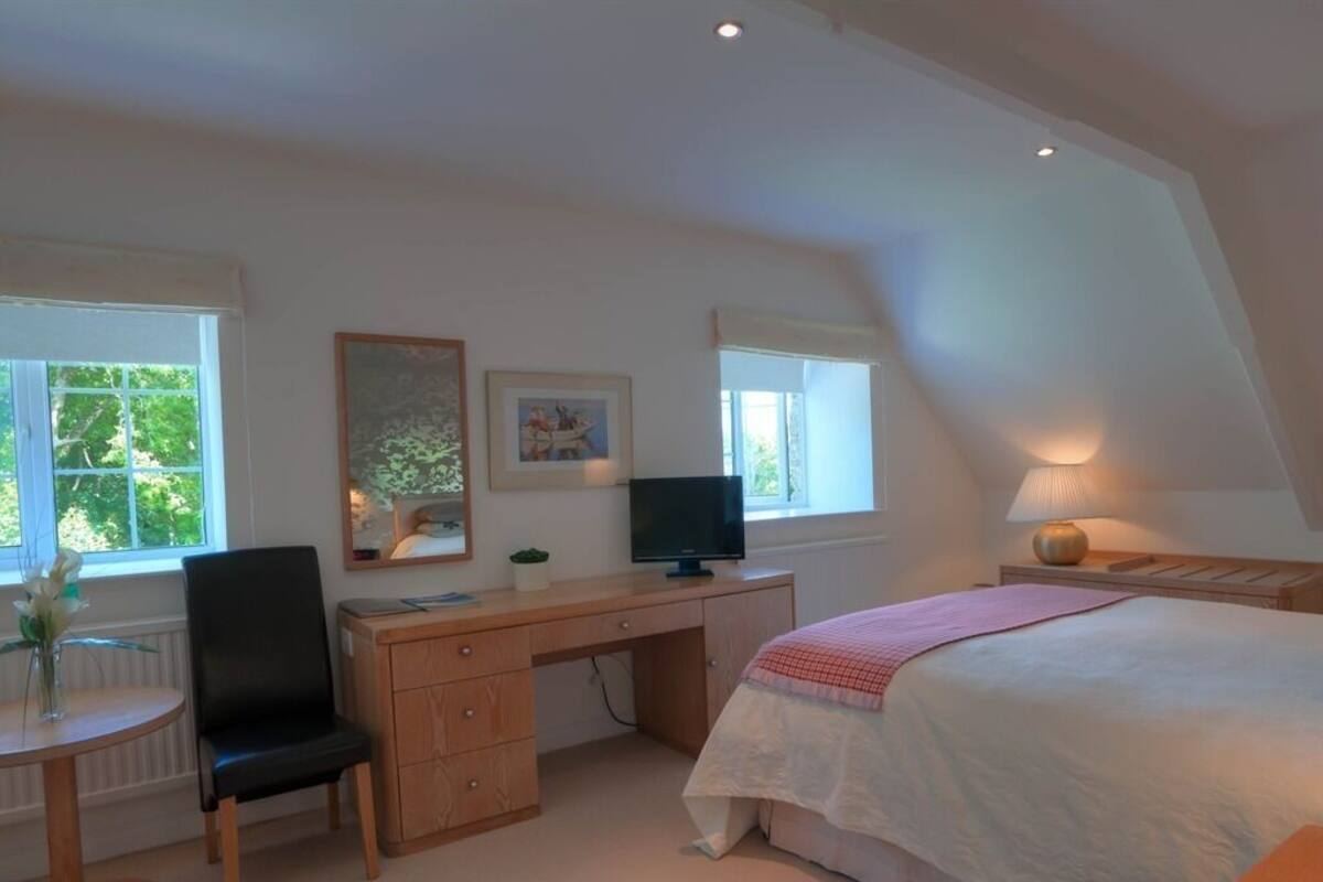 A bedroom with a bed, desk, chair, and a small table with a vase.