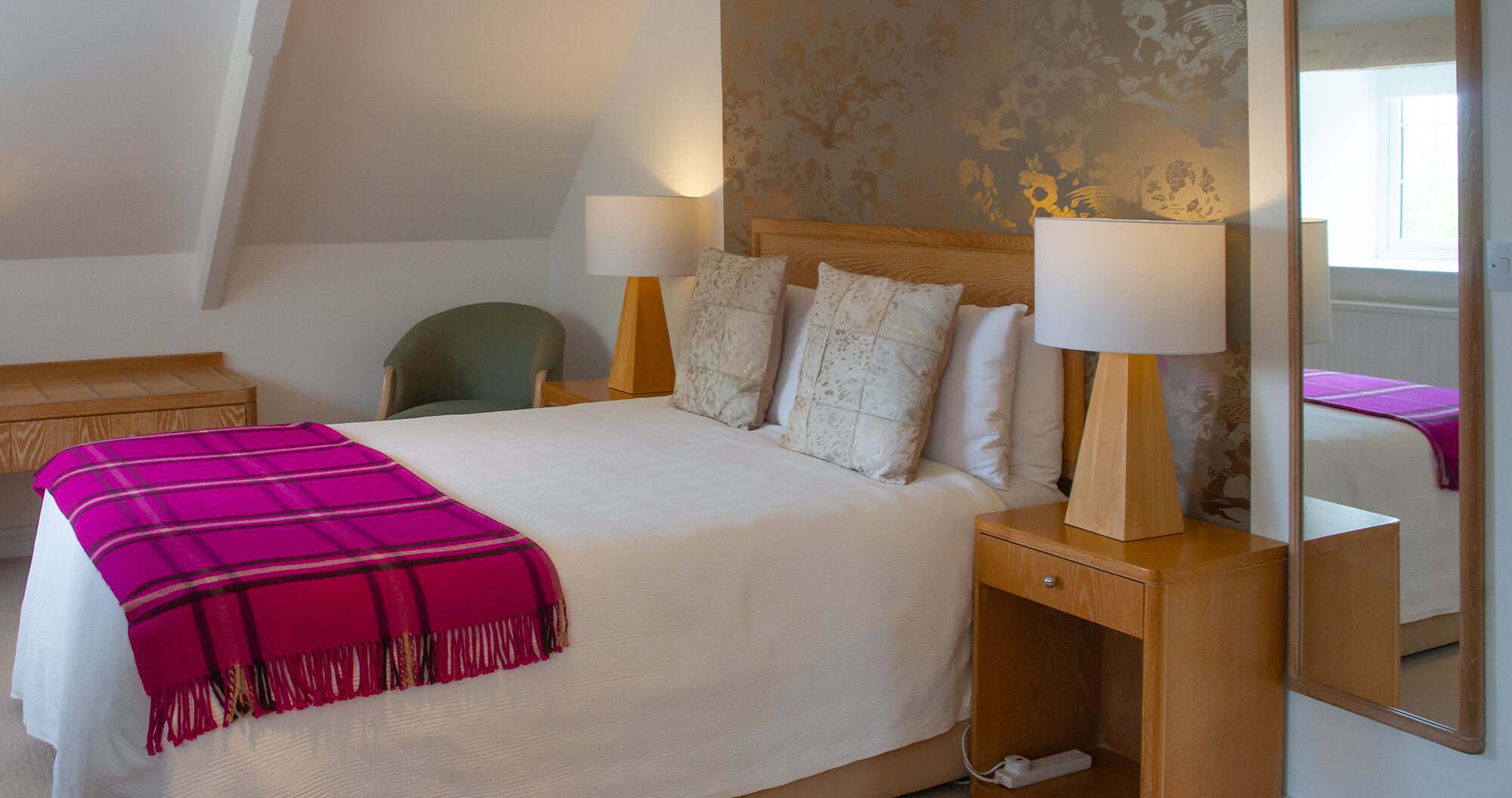 A hotel room with a bed, two bedside lamps, a wooden nightstand, a chair, a mirror, and a window.