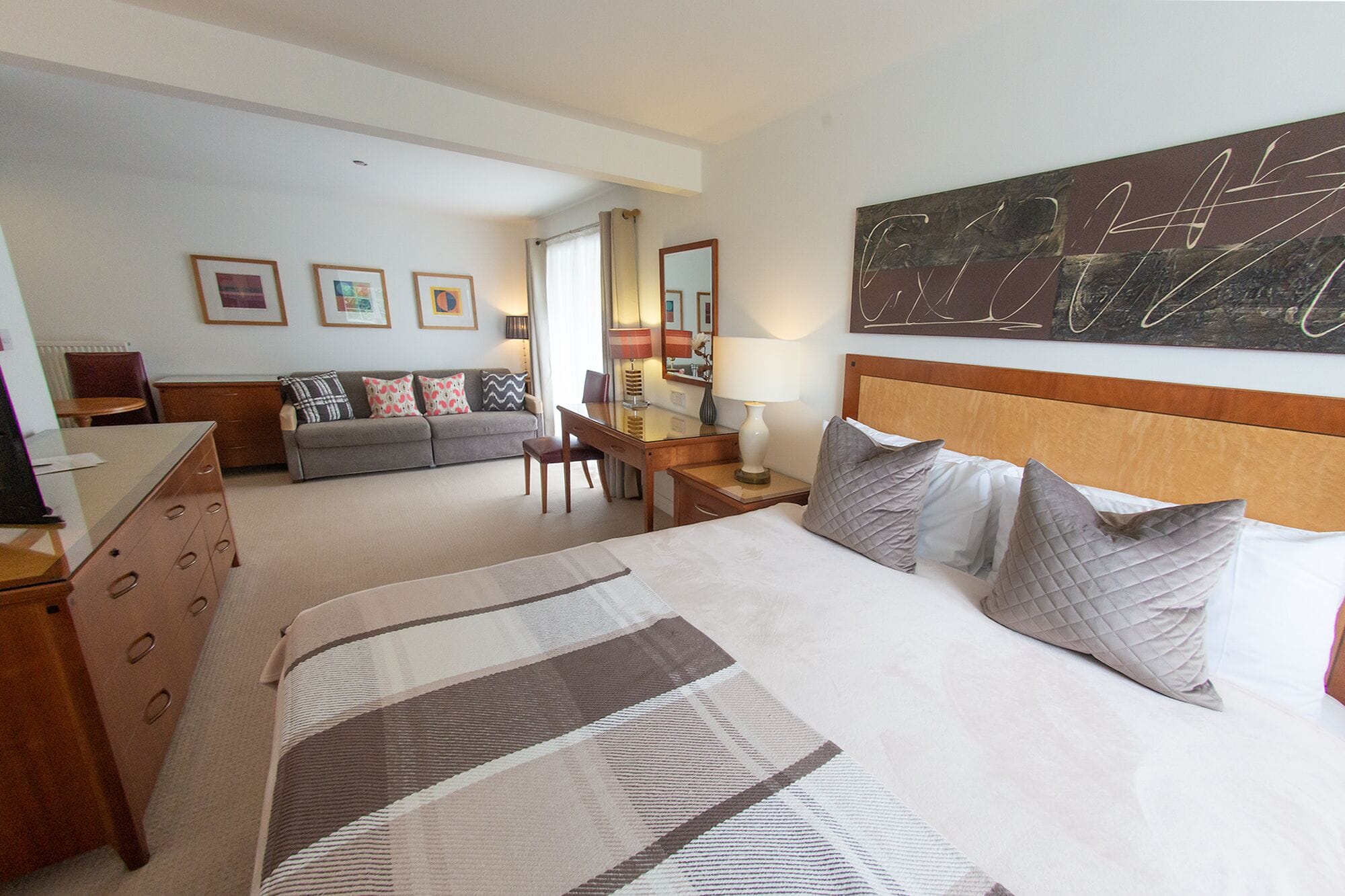 Habitación de hotel con una cama grande, un sofá, un escritorio y una silla. Se observan cuadros enmarcados en la pared y un cuadro abstracto de gran formato.