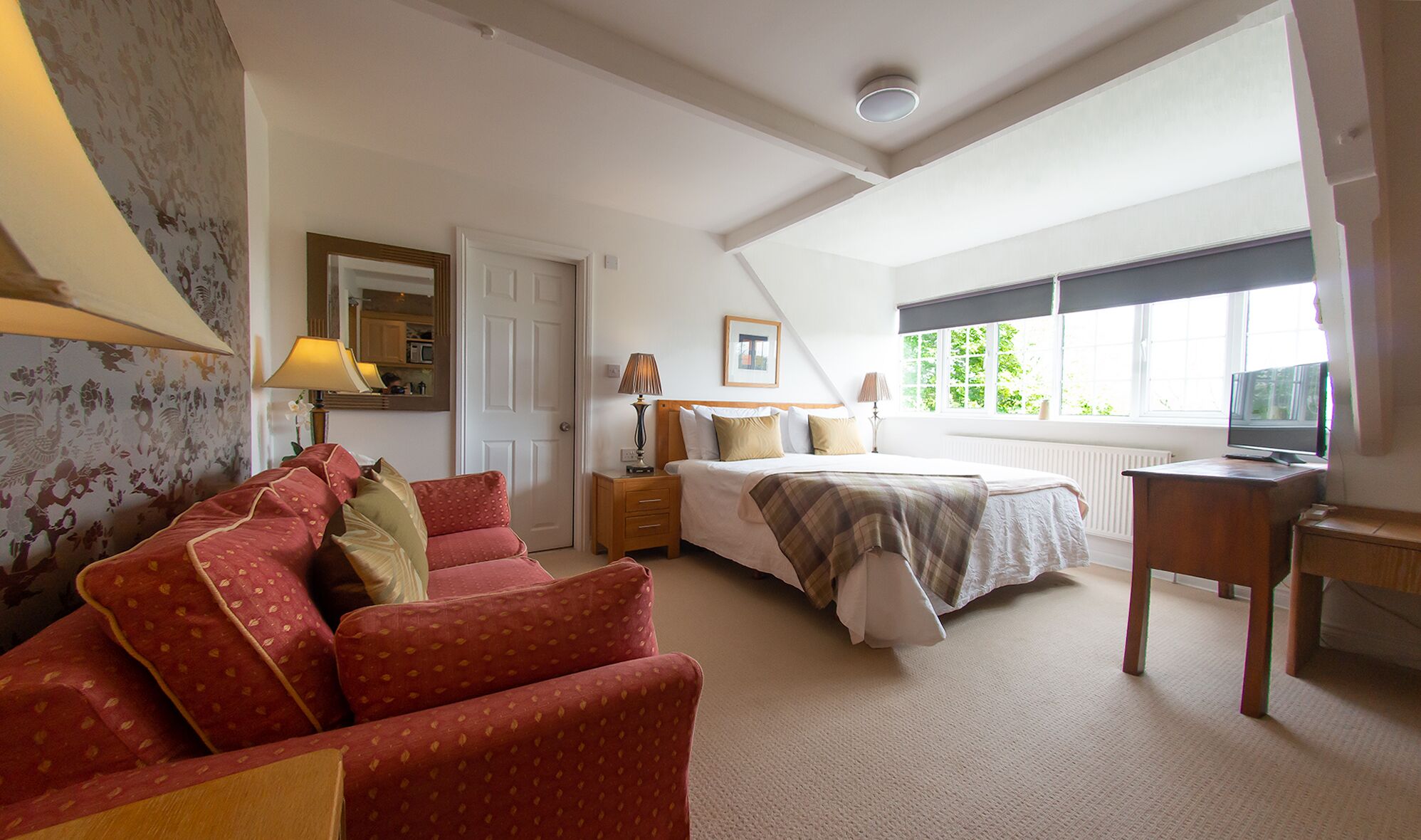 A bedroom with a bed, a desk, a chair, a nightstand, and a window with a view of greenery.
