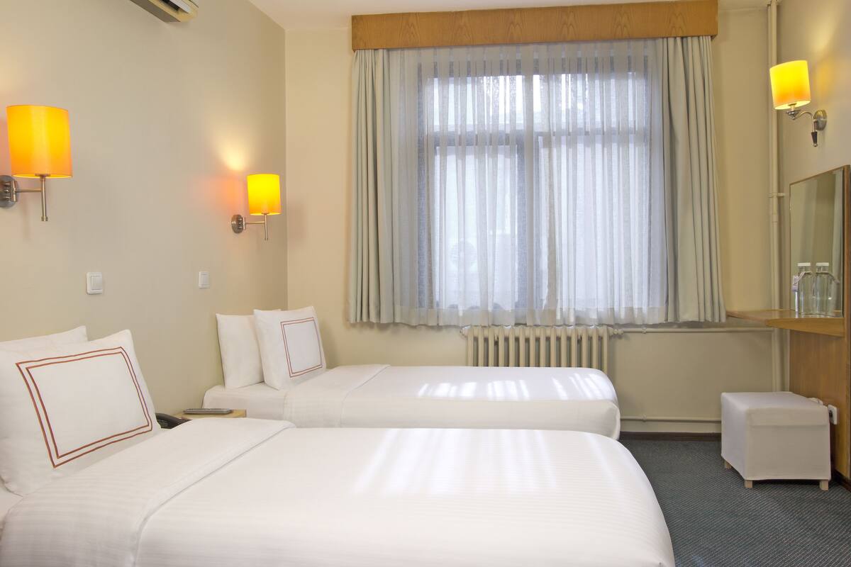 Hotellihuone, jossa on kaksi sänkyä, suuri ikkuna läpikuultavilla verhoilla, yöpöytä lampun kanssa, pieni jakkara ja peili seinällä.