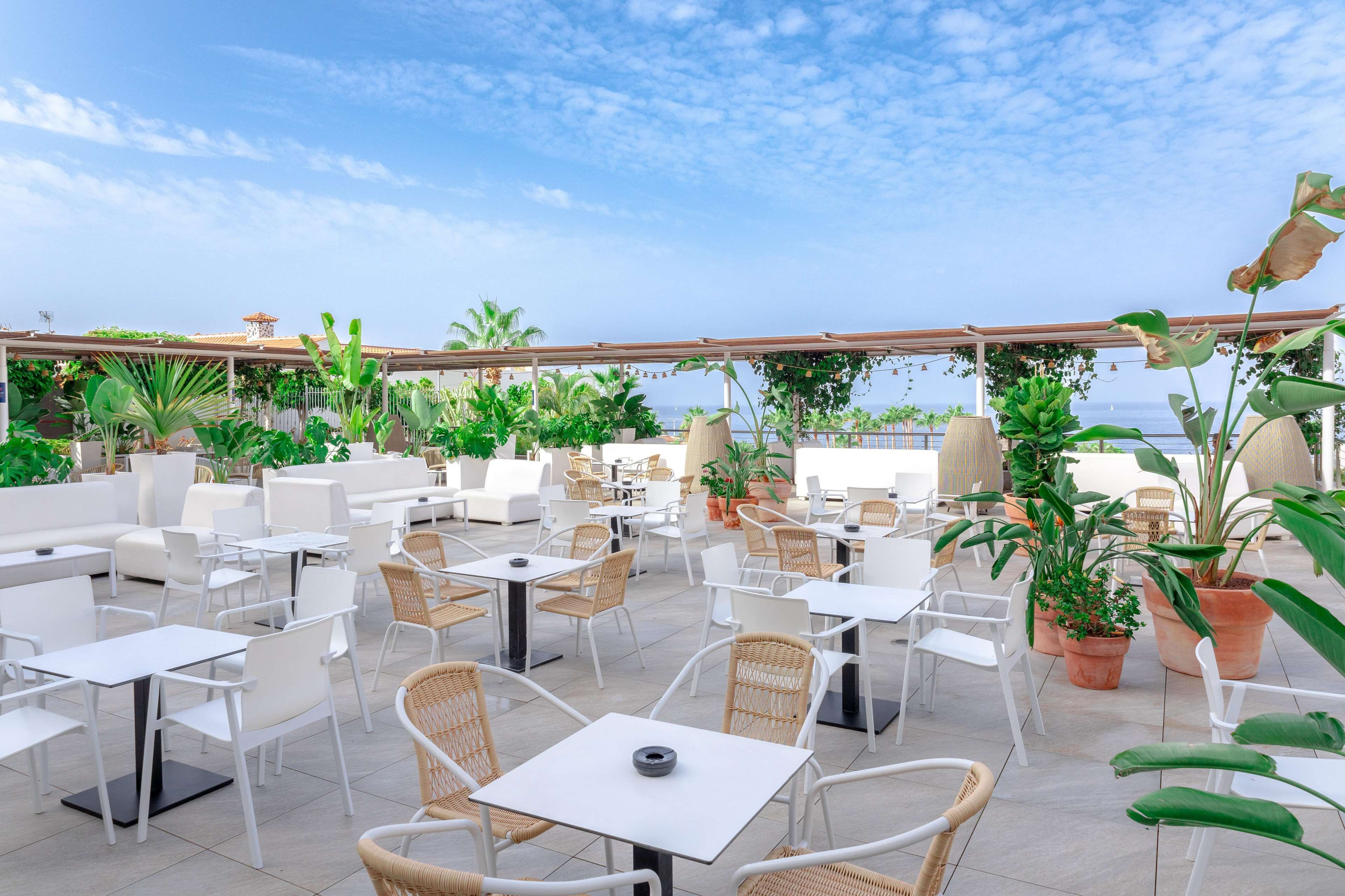 Een terras buiten met witte tafels en stoelen, potplanten en uitzicht op zee.