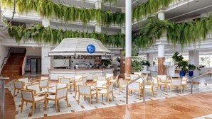 A spacious lobby with multiple seating areas, a central bar, and lush green plants hanging from the ceiling. - Landmar Playa La Arena (Santiago del Teide)