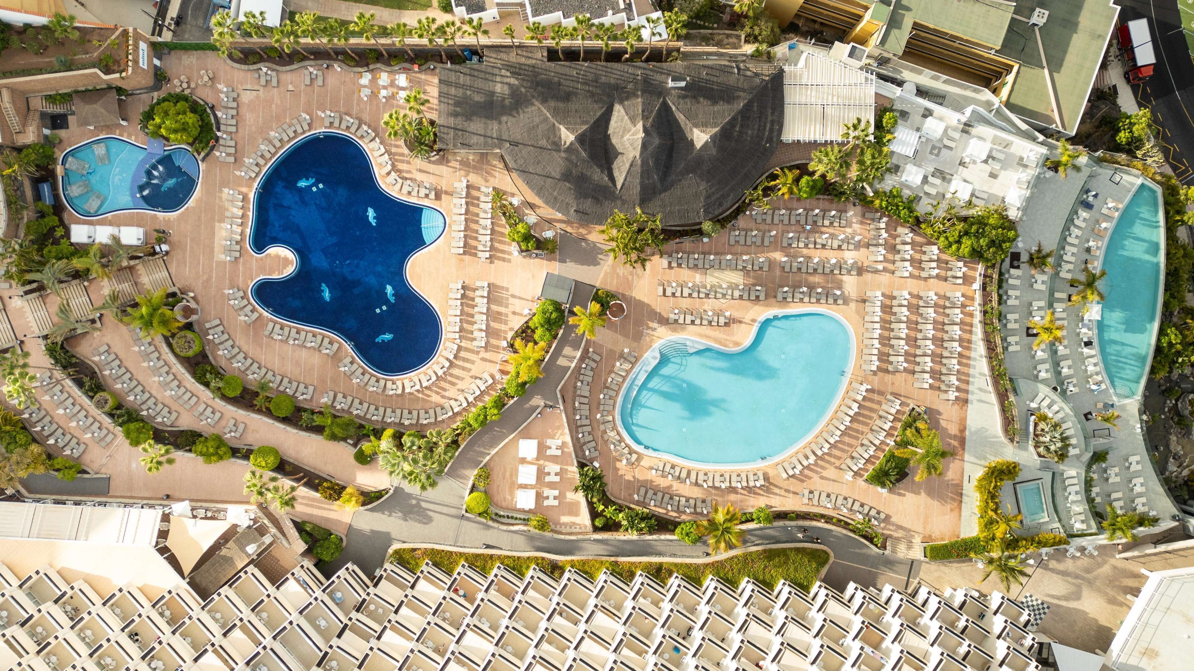 Vue aérienne d’un complexe hôtelier doté de plusieurs piscines, de chaises longues et d’un parking.