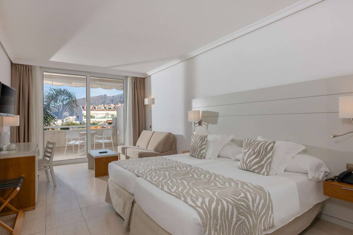 Ein Hotelzimmer mit einem großen Bett, einem Schreibtisch, einem Sessel, einem Fernseher und einem Balkon mit Bergblick.