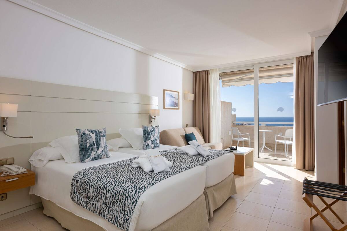 Ein Hotelzimmer mit zwei Betten, einem Balkon mit Meerblick, einem Schreibtisch und einem Fernseher.