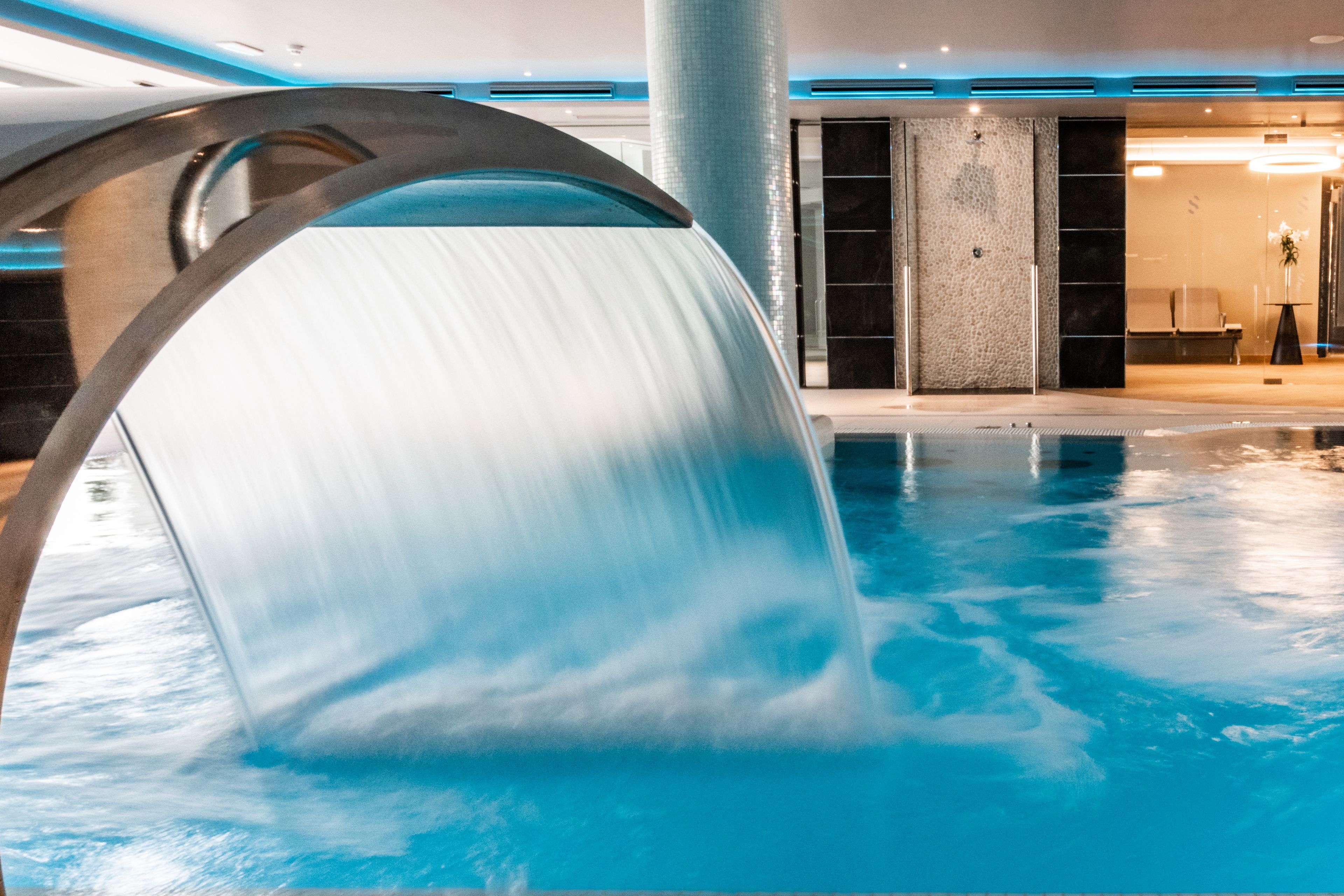 Una piscina interior moderna con un elemento acuático, una zona de recepción con un banco y una columna.