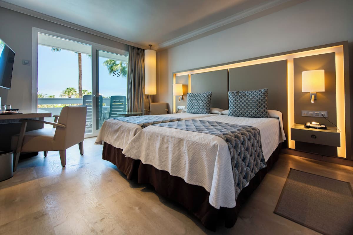 Hotelkamer met een bed, een bureau, een stoel, een televisie en een balkon met uitzicht op palmbomen.