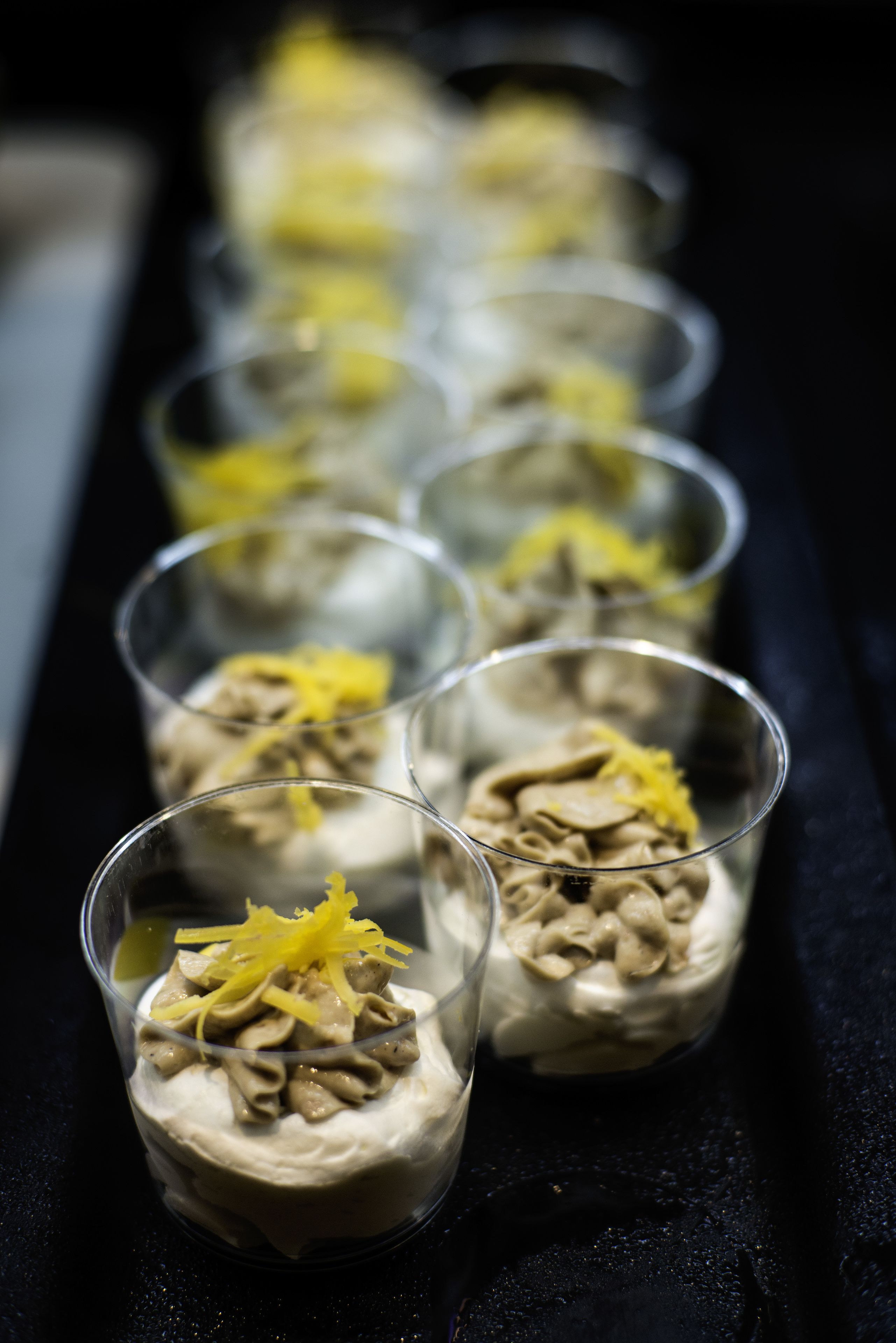 Une rangée de verres transparents contenant un dessert à base crémeuse, surmonté de ce qui ressemble à du fromage râpé ou à des zestes.