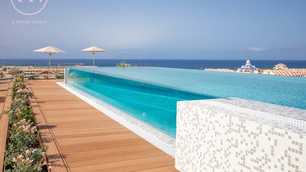 Una piscina infinita larga y cristalina con una terraza de madera y macizos de flores, con vistas a una playa y al océano.