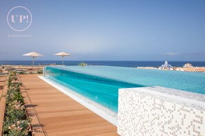 Una piscina infinita larga y cristalina con una terraza de madera y macizos de flores, con vistas a una playa y al océano.