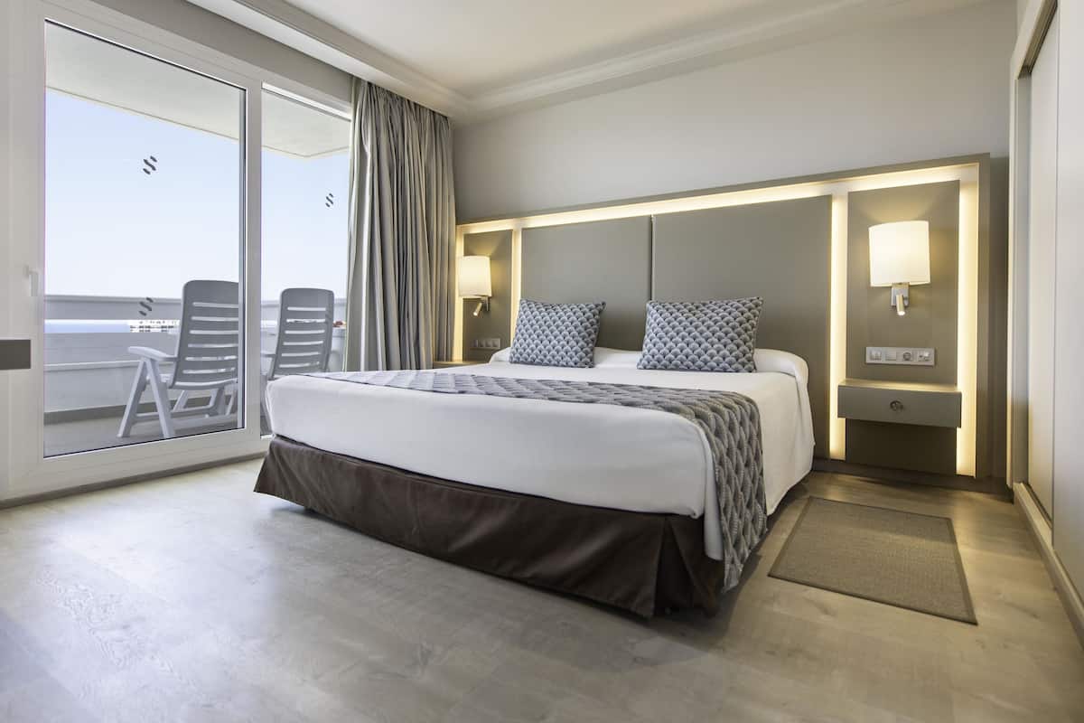 Een hotelkamer met een groot bed, een schuifglasdeur naar een balkon, een klein bureau en een nachtkastje met een lamp.