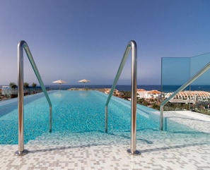 An infinity pool with metal railings, clear blue water, and a view of the ocean and buildings in the background.