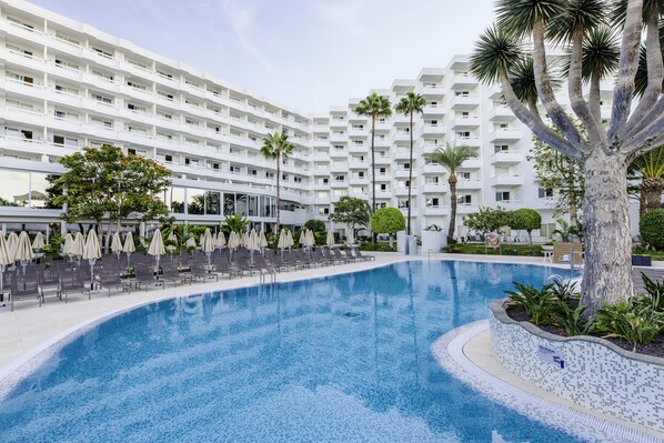 A hotel with a large pool surrounded by lounge chairs and umbrellas, palm trees, and a modern building with multiple balconies.