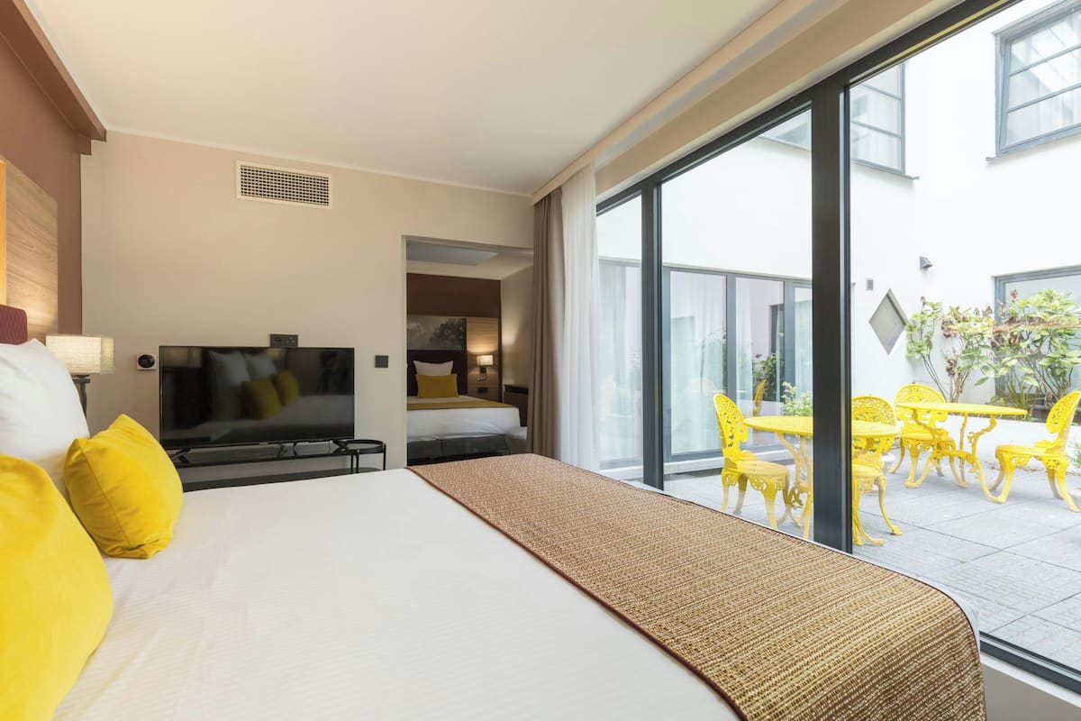 A hotel room with a bed, television, and a view of an outdoor patio with yellow chairs and tables.