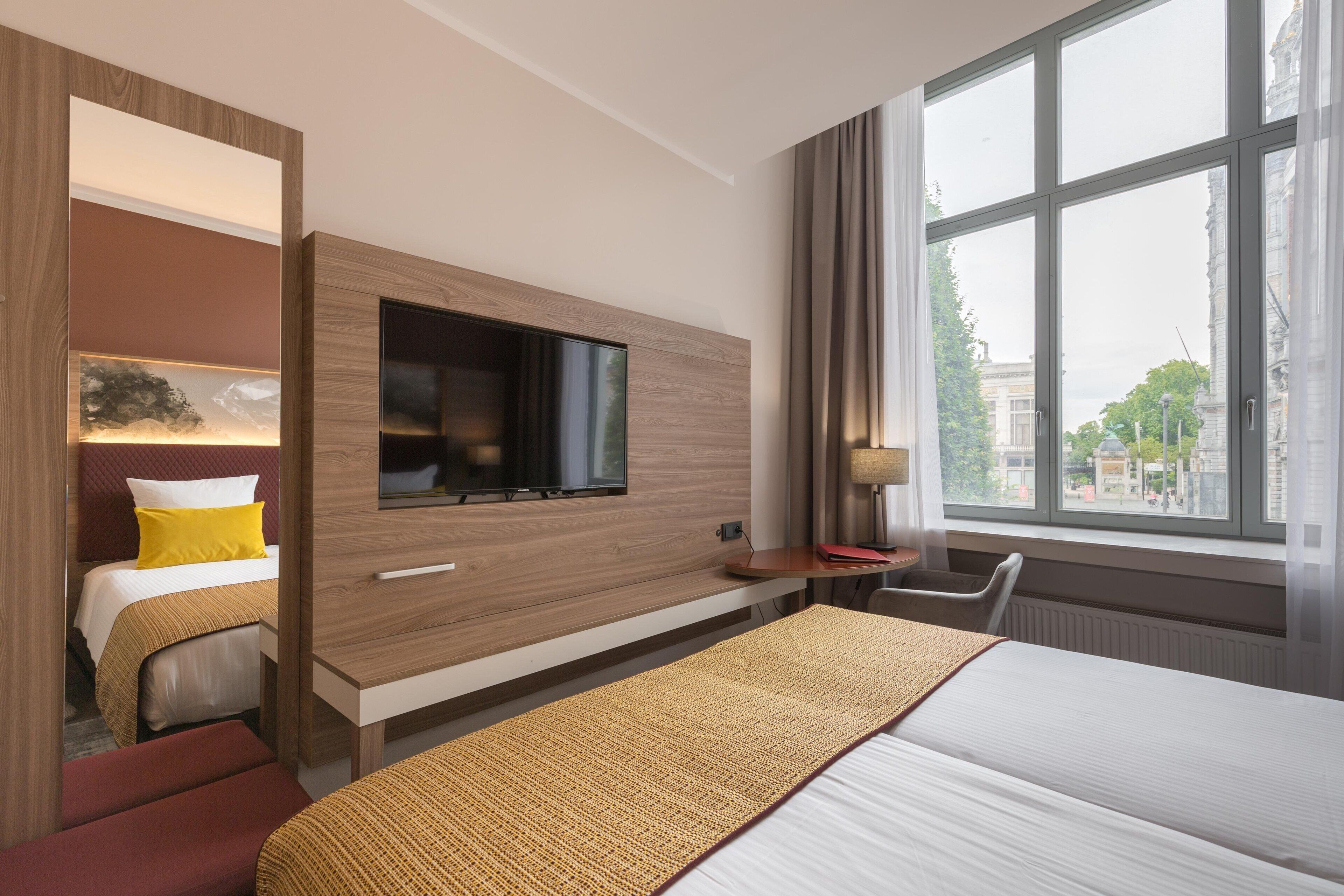 A hotel room with a bed, a TV mounted on the wall, a desk, and a window with a view of buildings.