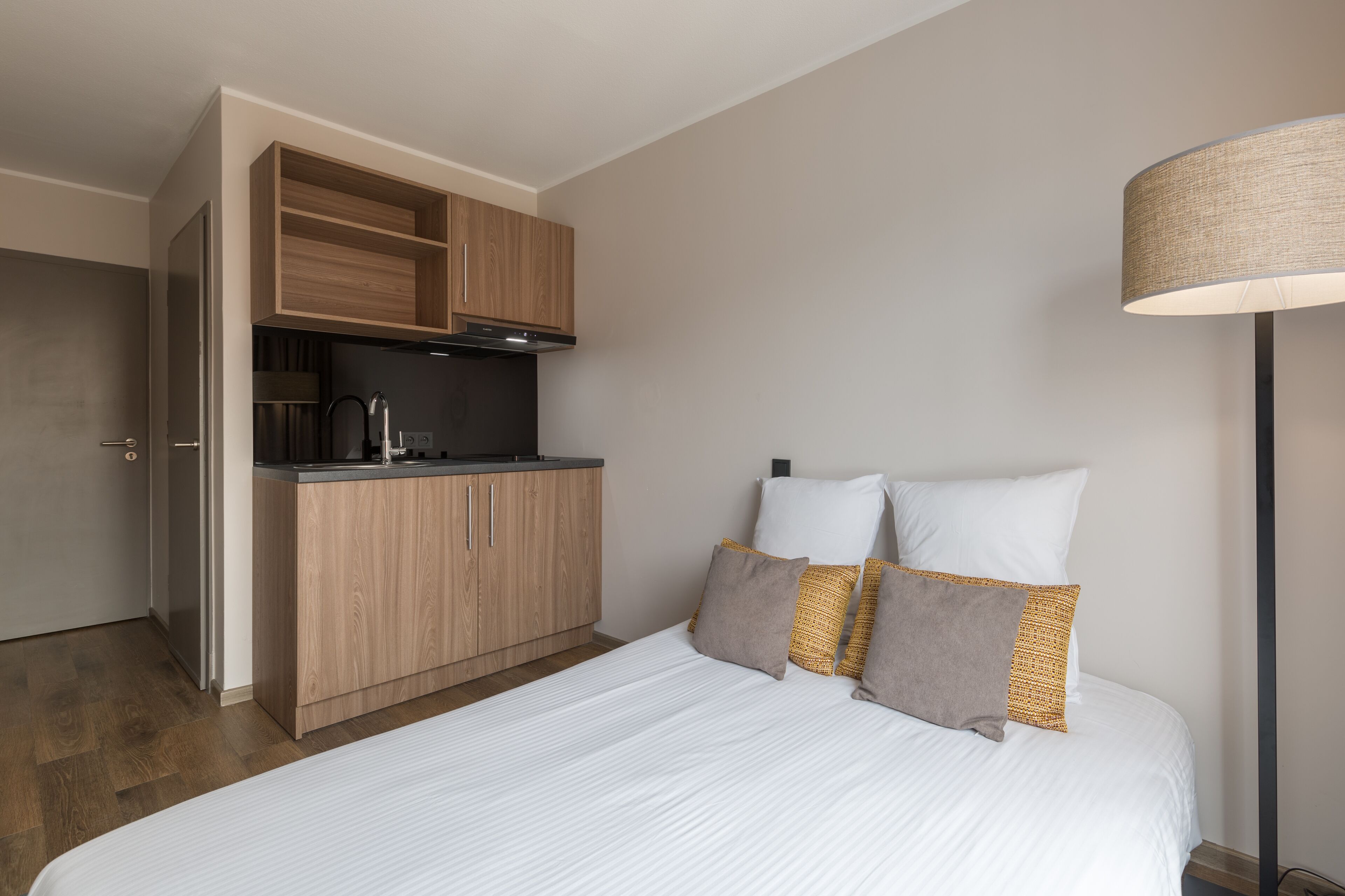 A compact hotel room with a kitchenette, a bed with white bedding, and a floor lamp.