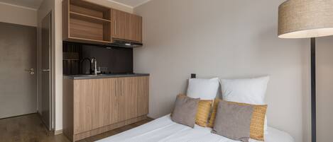 A compact hotel room with a kitchenette, a bed with white bedding, and a floor lamp.