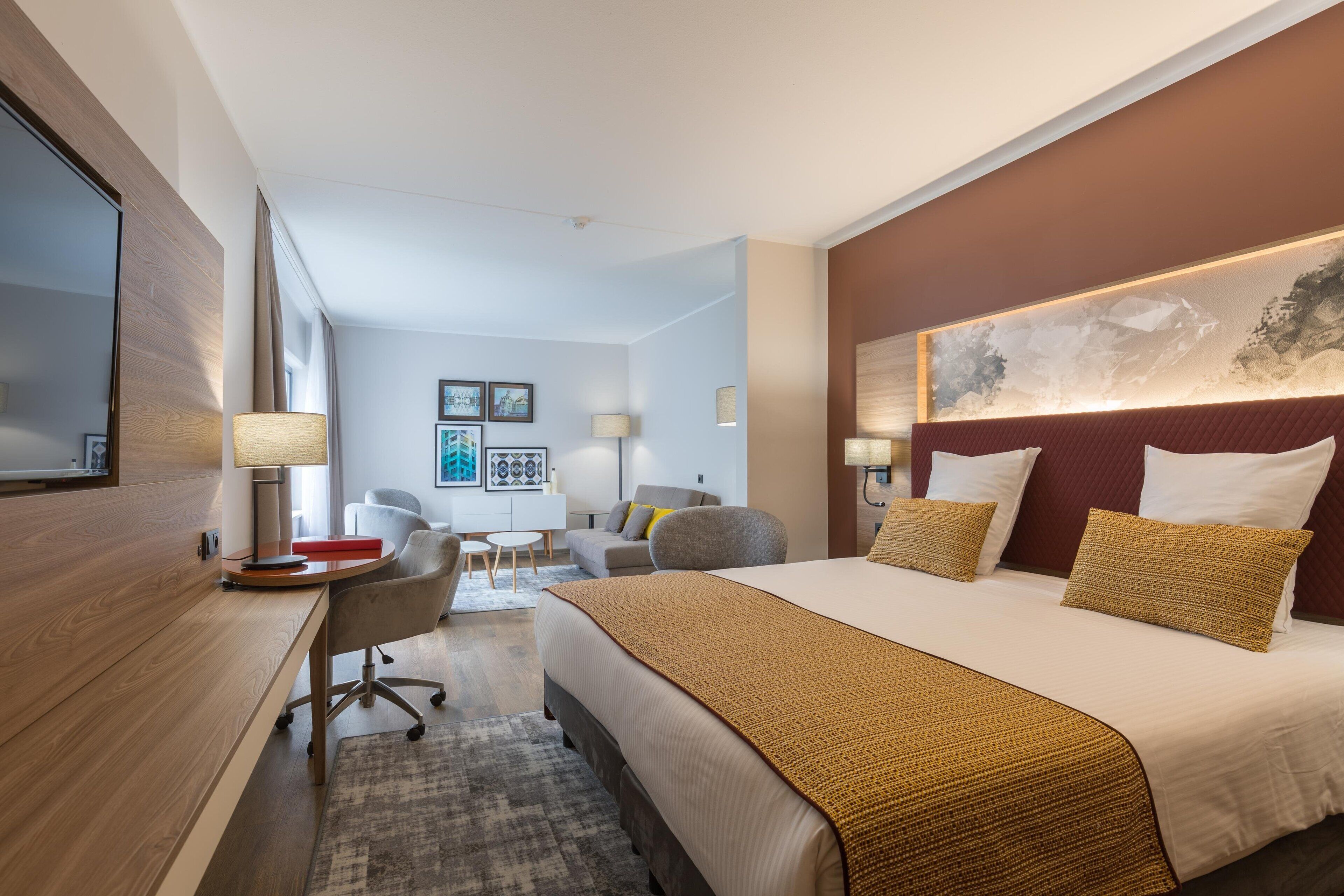 A modern hotel room with a large bed, a desk, and a chair. There is a painting on the wall and a window with curtains.