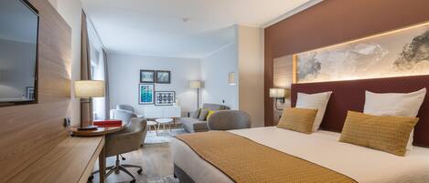 A modern hotel room with a large bed, a desk, and a chair. There is a painting on the wall and a window with curtains.