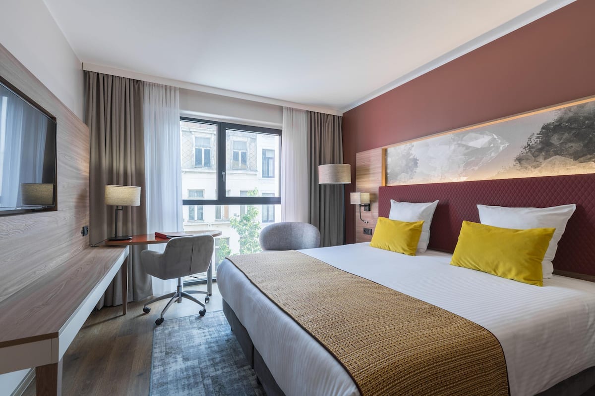 A hotel room with a bed, desk, chair, and large windows.