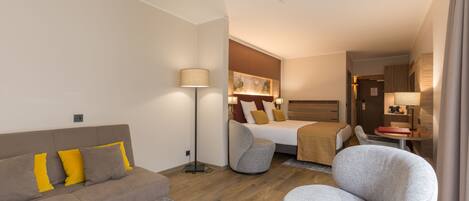 A modern hotel room with a sofa, armchair, and a bed with a headboard.