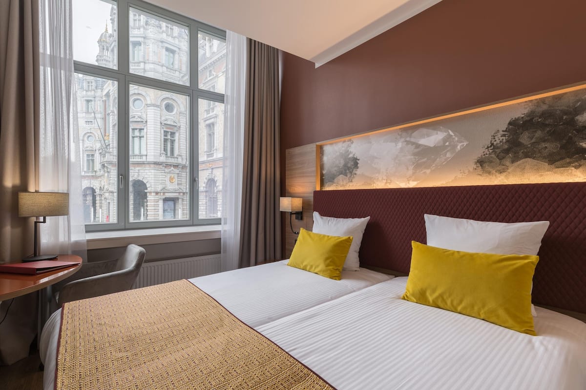 Una habitación de hotel con un ventanal grande, una cama con almohadas blancas y amarillas, un escritorio con lámpara y una obra de arte colgada en la pared.