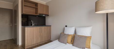 A compact hotel room with a kitchenette, a bed with white bedding, and a floor lamp.