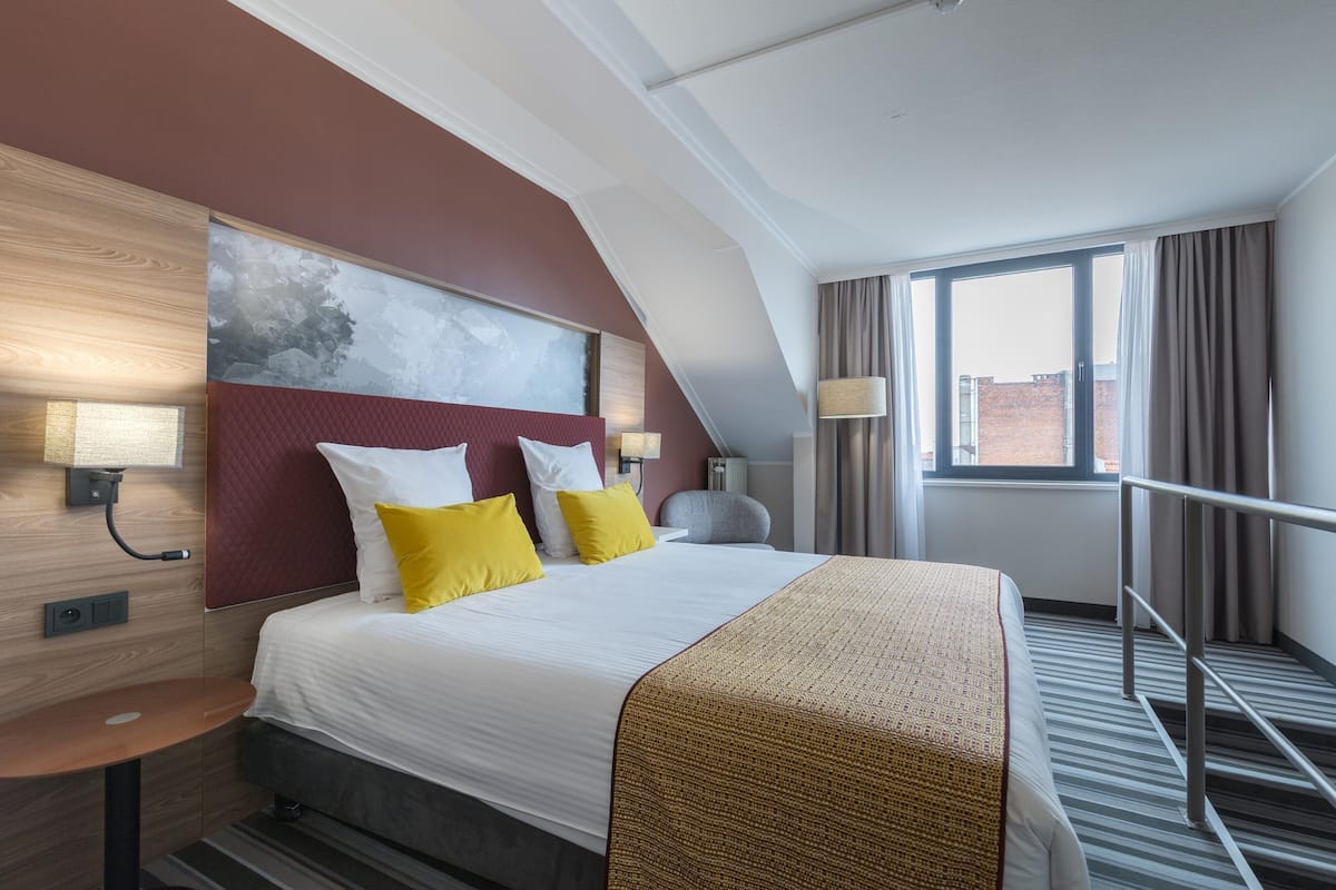 A hotel room with a large bed, two bedside lamps, a headboard with a painting, a window with curtains, and a view of buildings outside.
