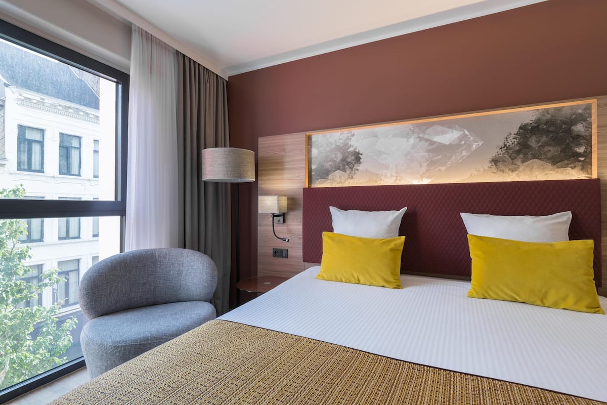 Habitación de hotel con una cama grande, dos almohadas, una mesita de noche y una lámpara, con vista a un edificio a través de una ventana.