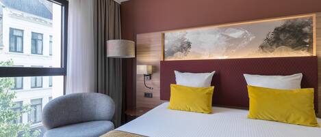 A hotel room with a large bed, two pillows, a side table, a lamp, and a view of a building through a window.