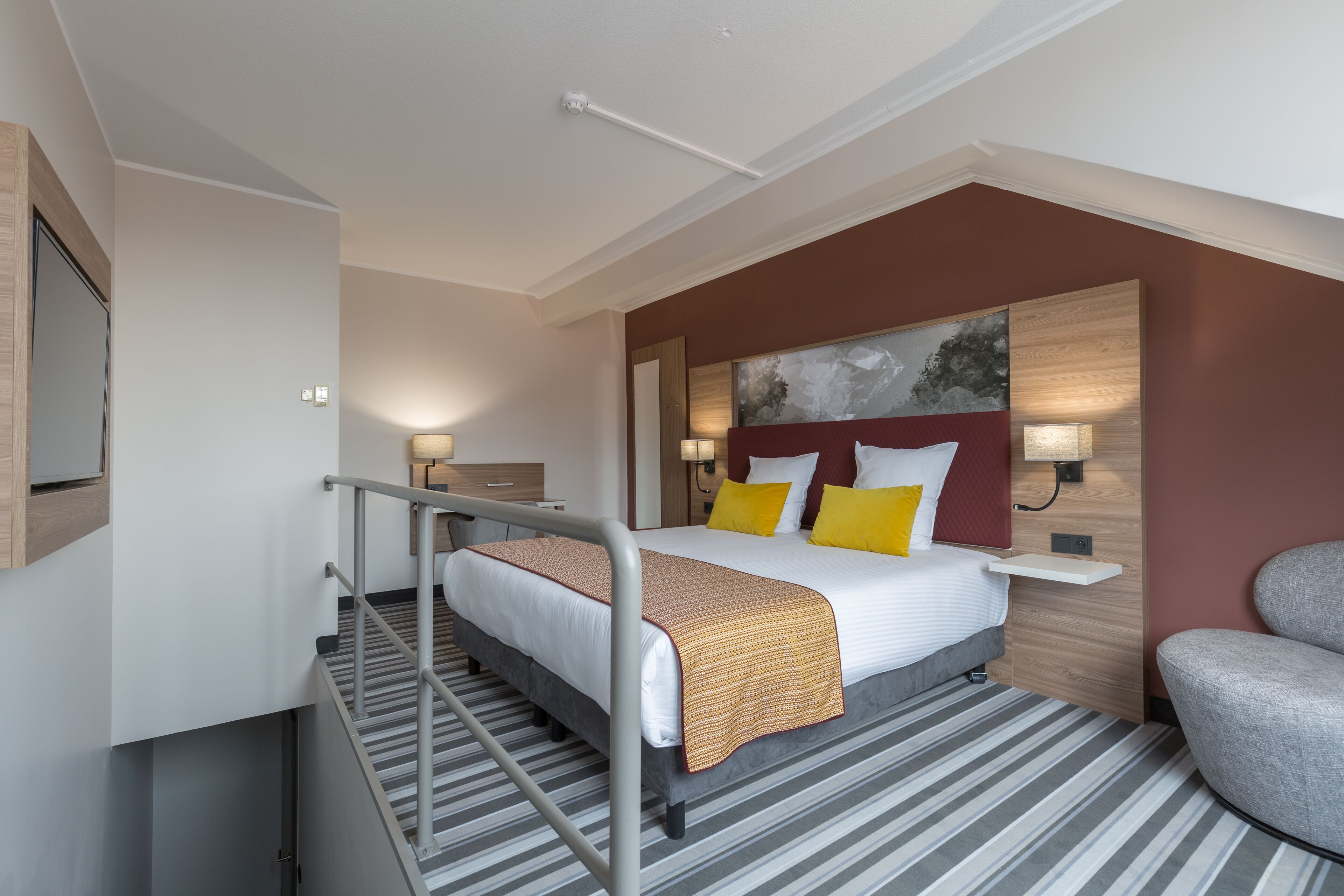 A hotel room with a bed, a chair, a desk, and a painting on the wall.