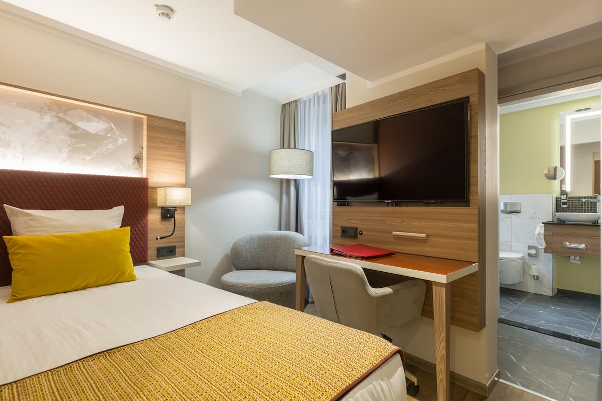 A hotel room with a bed, a desk, a chair, a television, a lamp, and a bathroom visible through an open door.