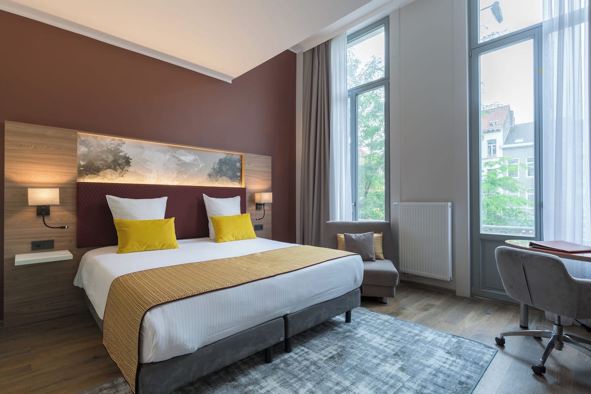 A modern hotel room with a large bed, a desk, and a view of the city through the window.