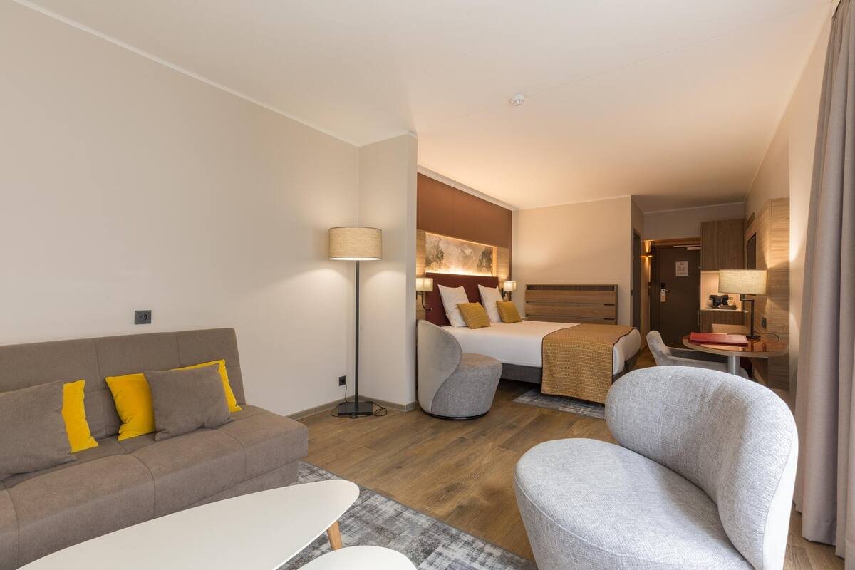 A modern hotel room with a sofa, armchair, and a bed with a headboard.