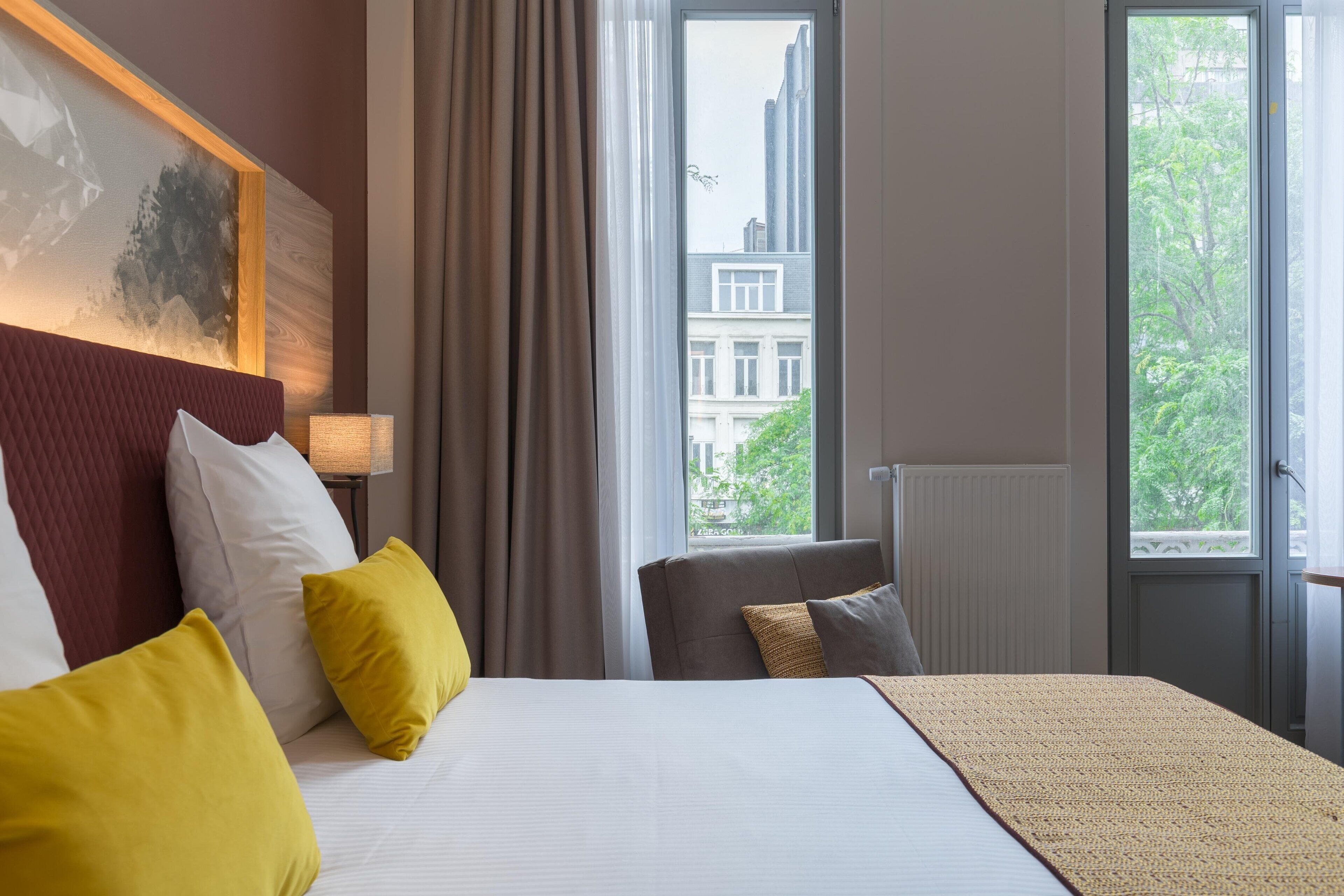 A hotel room with a large bed, a view of buildings through the window, and a chair with cushions.