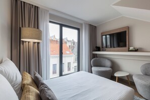 A hotel room with a bed, a desk with a flat-screen TV, a chair, a small table, and a view of buildings through the window. - Leonardo Hotel Antwerp (Antwerp)