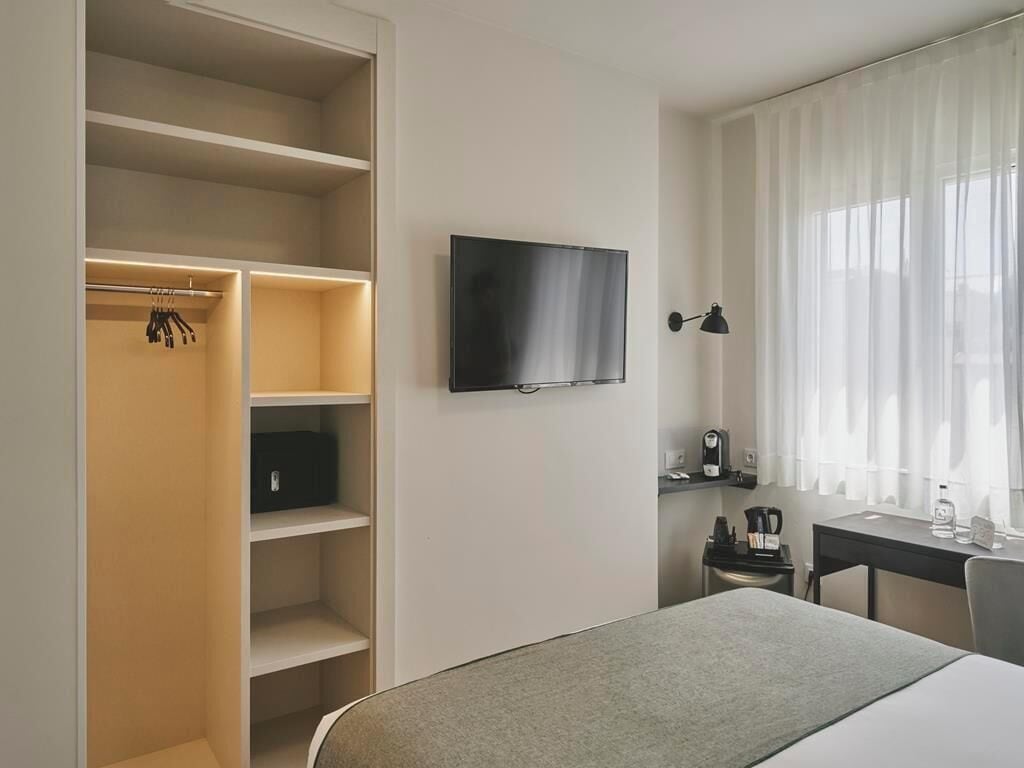 A hotel room with a bed, a TV mounted on the wall, a desk with a chair, a coffee machine, and a wardrobe with shelves.