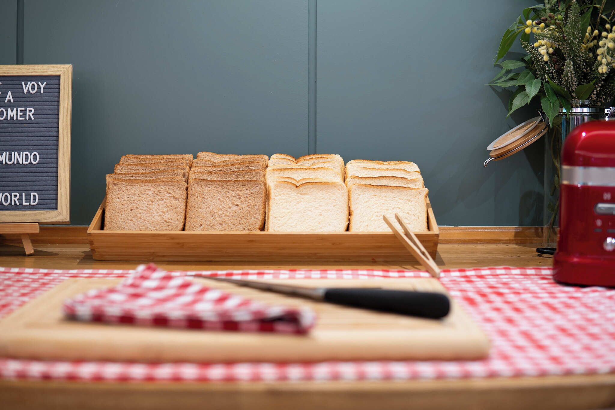 A wooden bread box with sliced bread on a red and white checkered tablecloth, a knife, and a red appliance on a table.