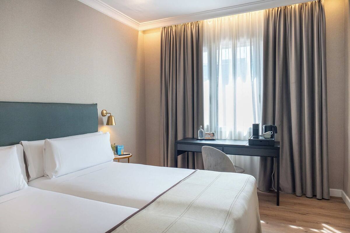 Une chambre d’hôtel avec un lit, un bureau, une chaise et une fenêtre avec des rideaux.