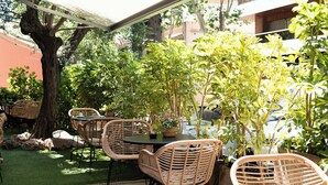An outdoor patio with wicker chairs and tables, surrounded by greenery and a tree. - Tres Torres Atiram Hotel (Barcelona)