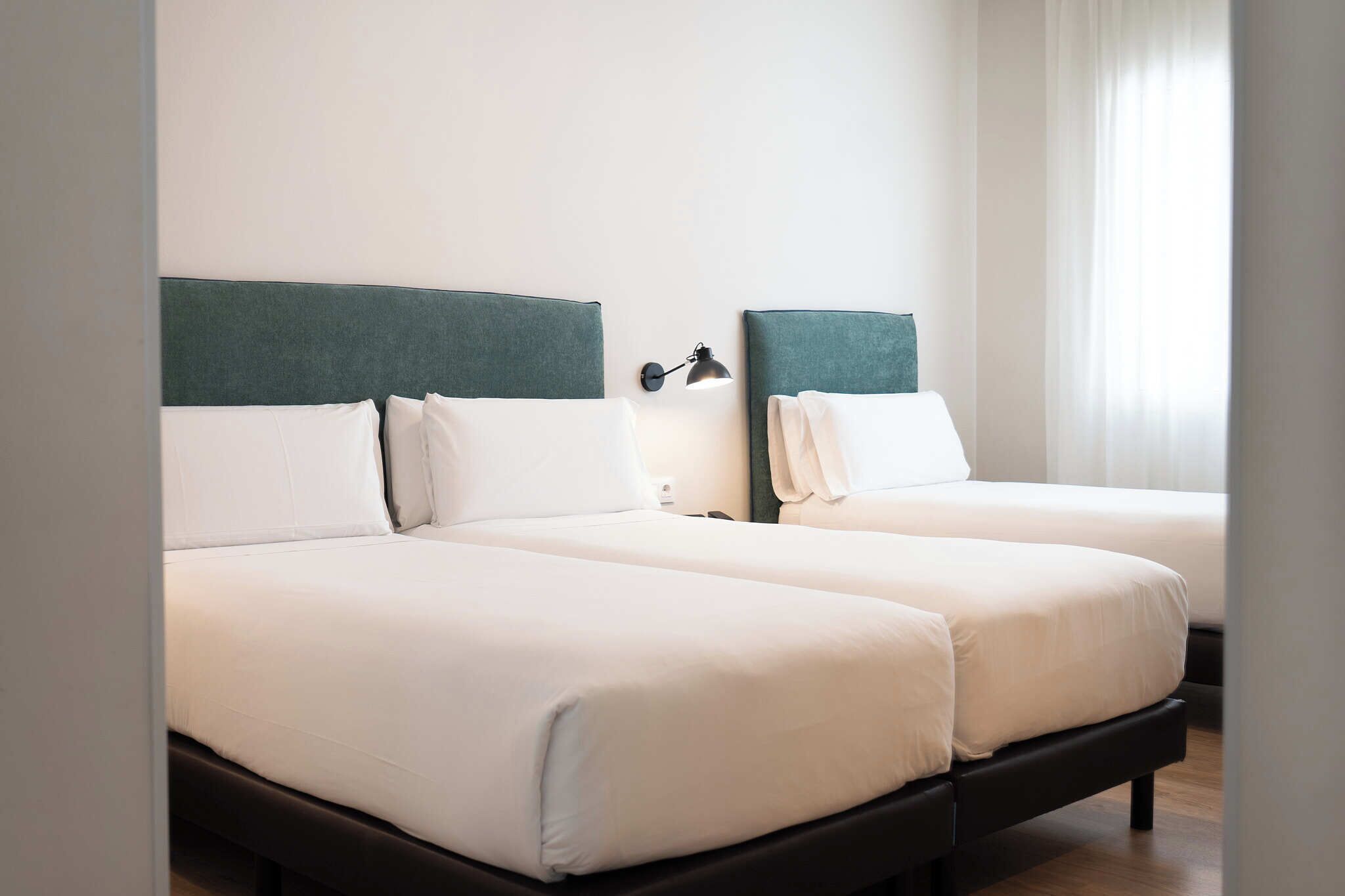 Dos camas individuales con ropa blanca y cabeceros oscuros, una lámpara de pared y una ventana con cortinas de gasa.
