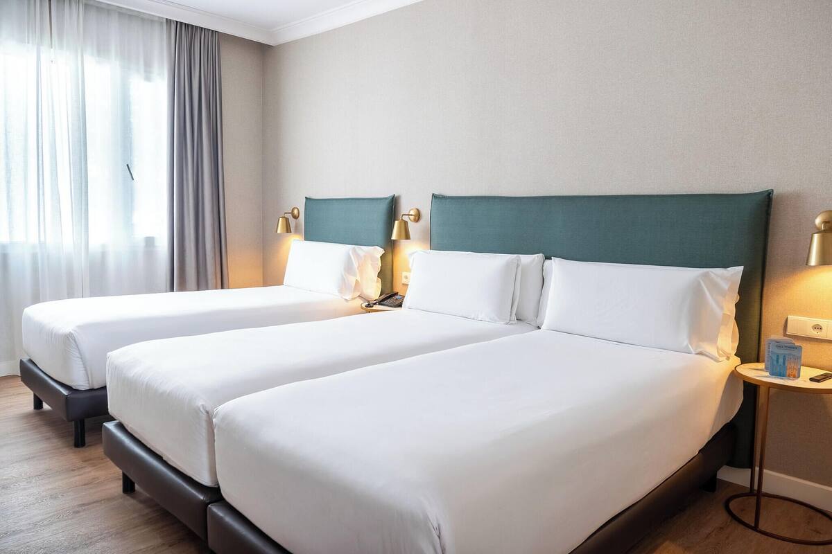 Une chambre d’hôtel avec deux lits, chacun doté de draps blancs et d’une tête de lit verte. On y trouve des lampes de chevet et une petite table sur laquelle sont posés un livre et un téléphone.