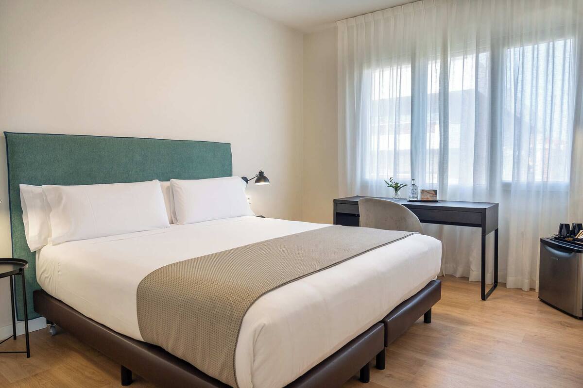 Un lit bien fait, avec une tête de lit verte, une table de chevet avec une lampe et un bureau avec une chaise.