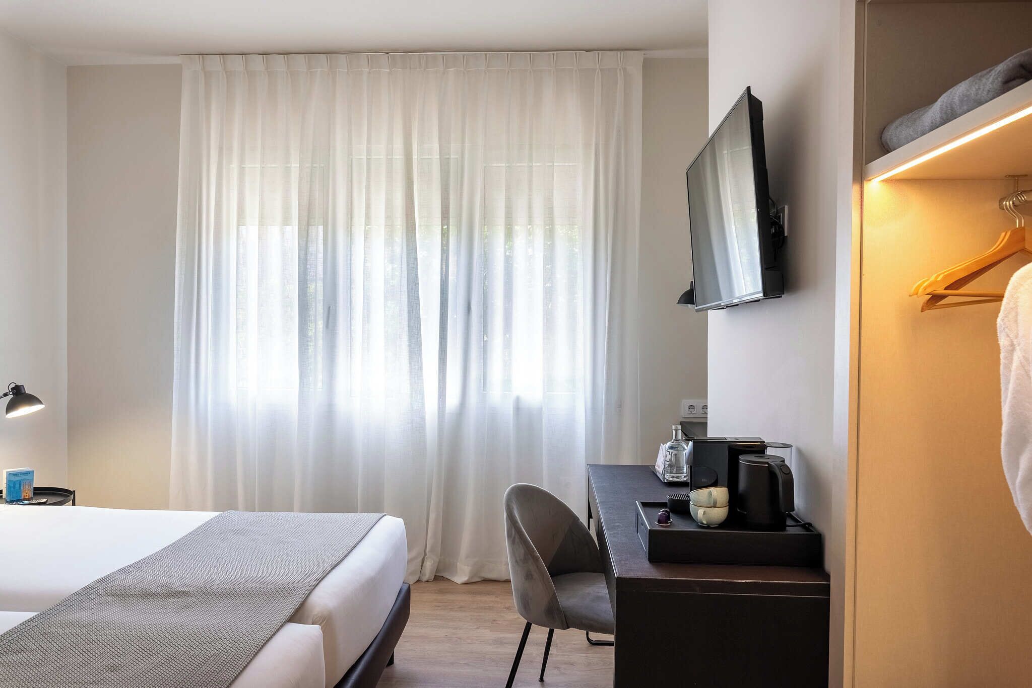 Une chambre d’hôtel comprenant un lit, un bureau avec une machine à café, une chaise, un téléviseur fixé au mur et une fenêtre avec des rideaux légers.