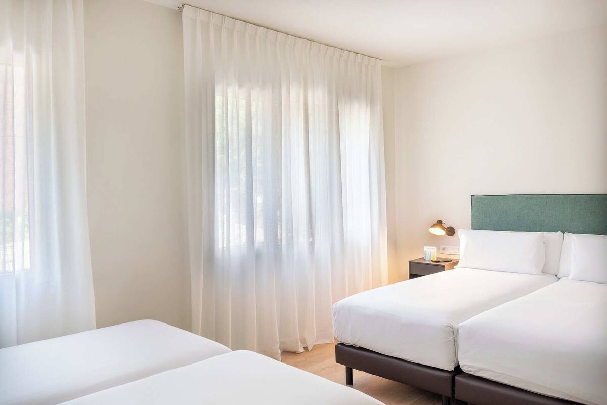 Une chambre d’hôtel avec deux lits, une table de chevet avec une lampe et des rideaux légers.