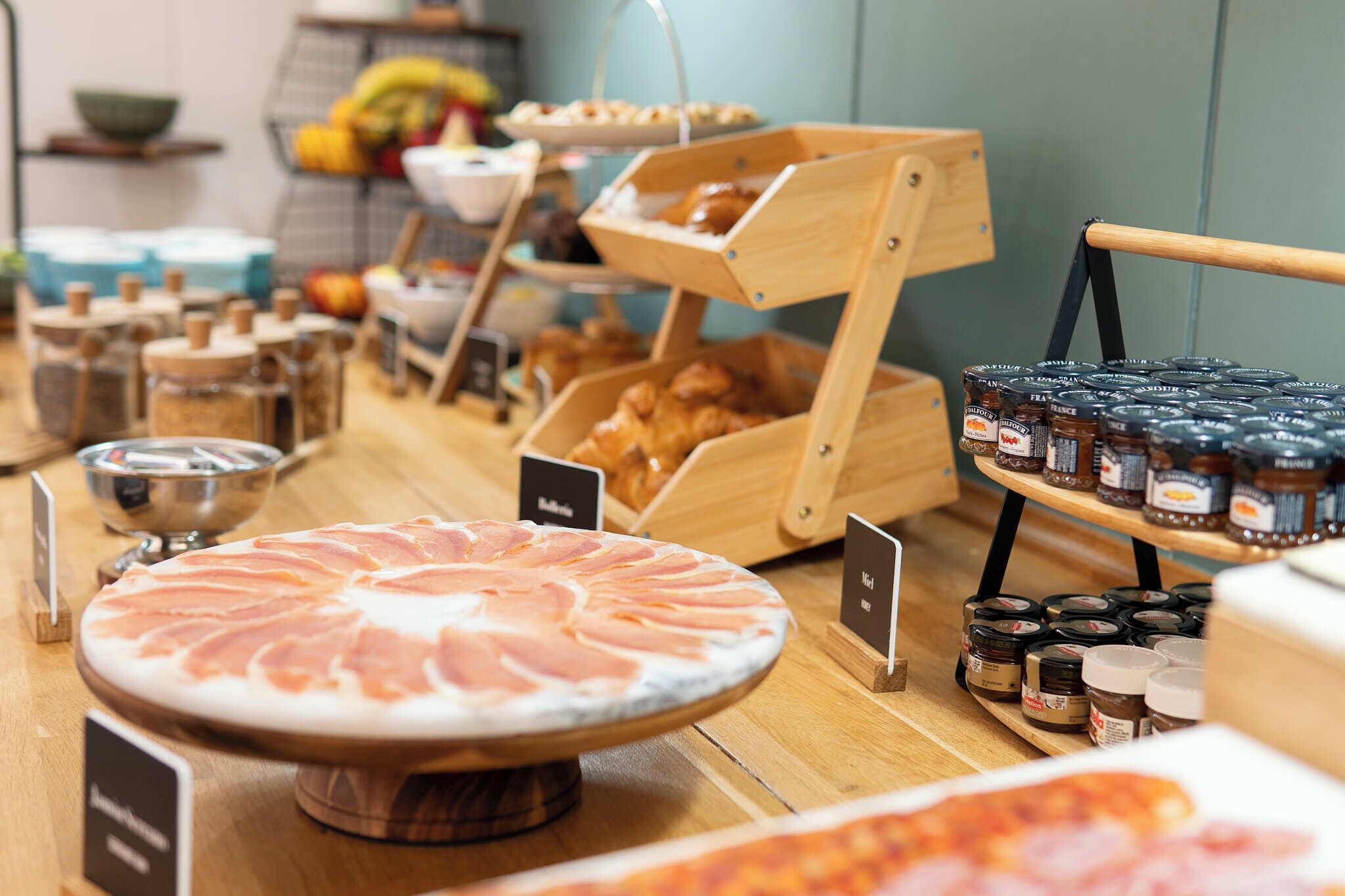Un comptoir de produits alimentaires proposant une variété d’articles, notamment un grand plateau de jambon de Parme, des pots de condiments et un assortiment de pains.